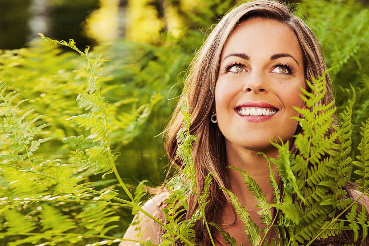 A woman outdoors with her head peeking out a bush as she smiles looking up.